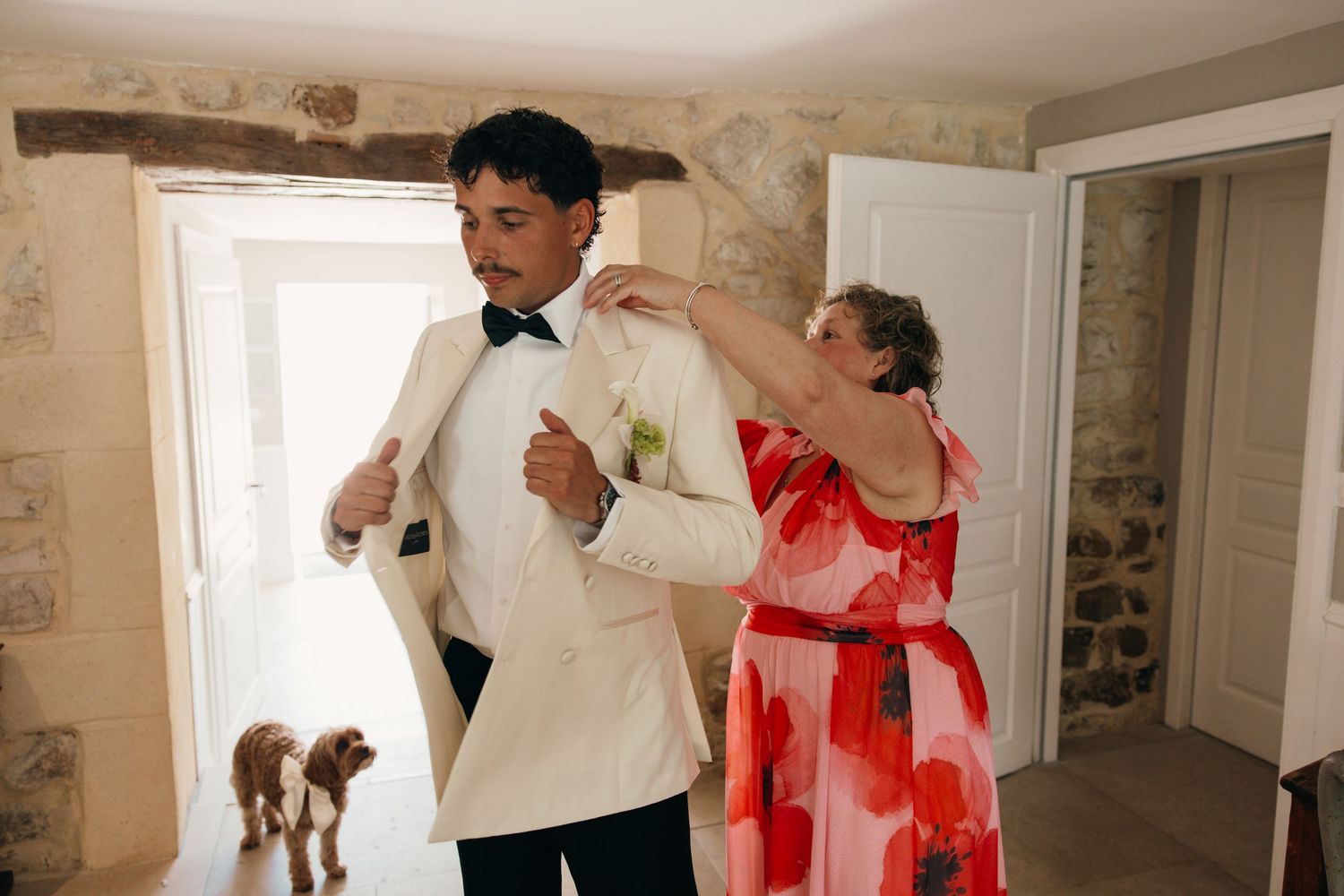 Groom in white tuxedo being helped with jacket by woman in floral dress with dog nearby.