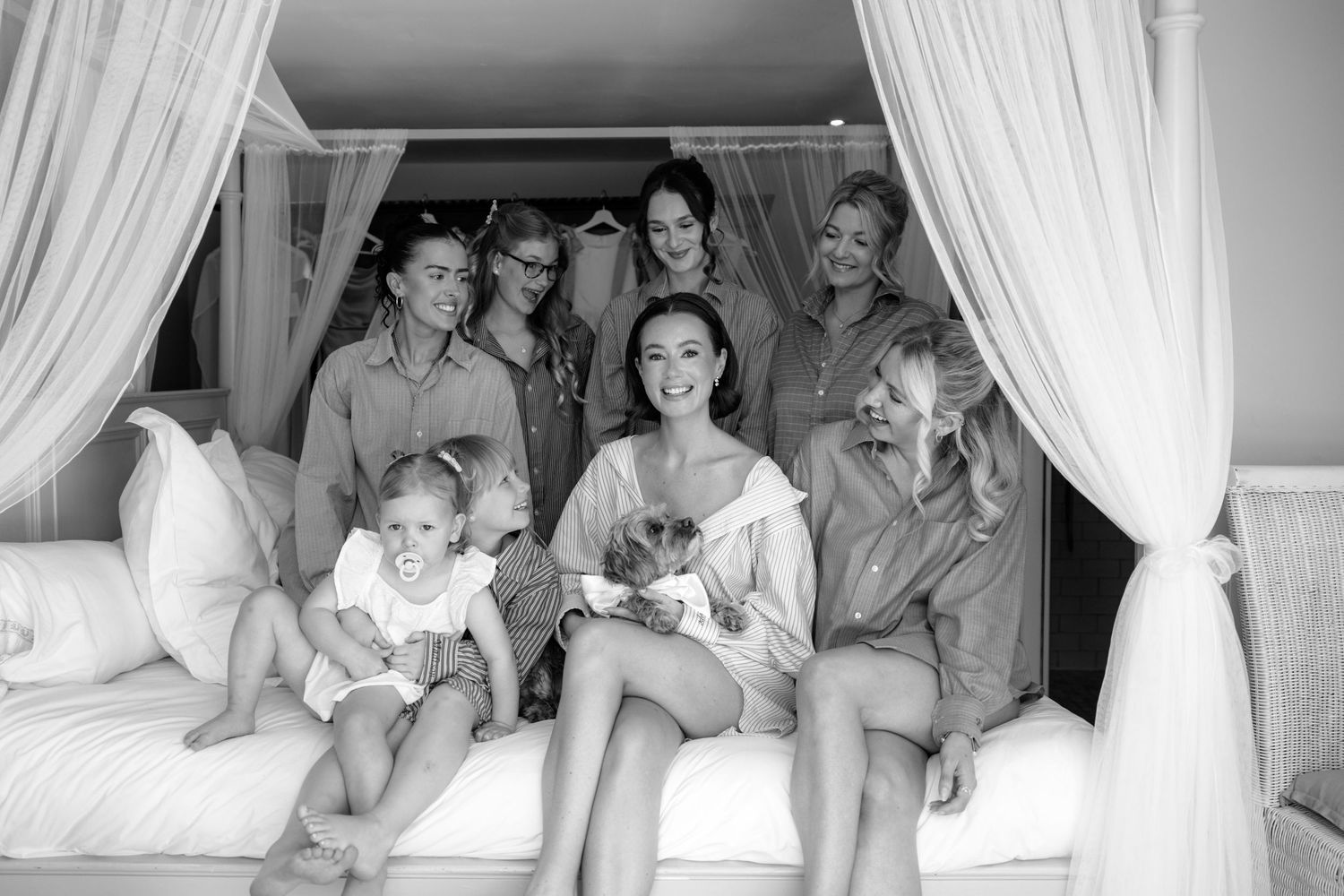 Bride poses with bridesmaids and a small child on a canopy bed in black and white photo.