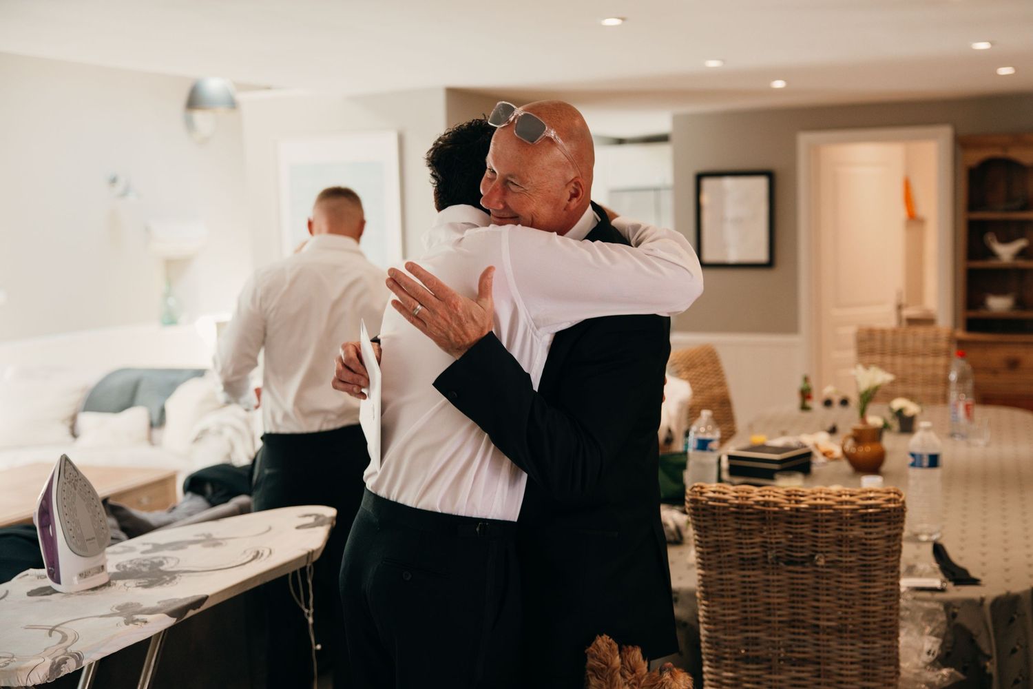 Two people share an emotional hug indoors during wedding preparations, with another guest visible in background.