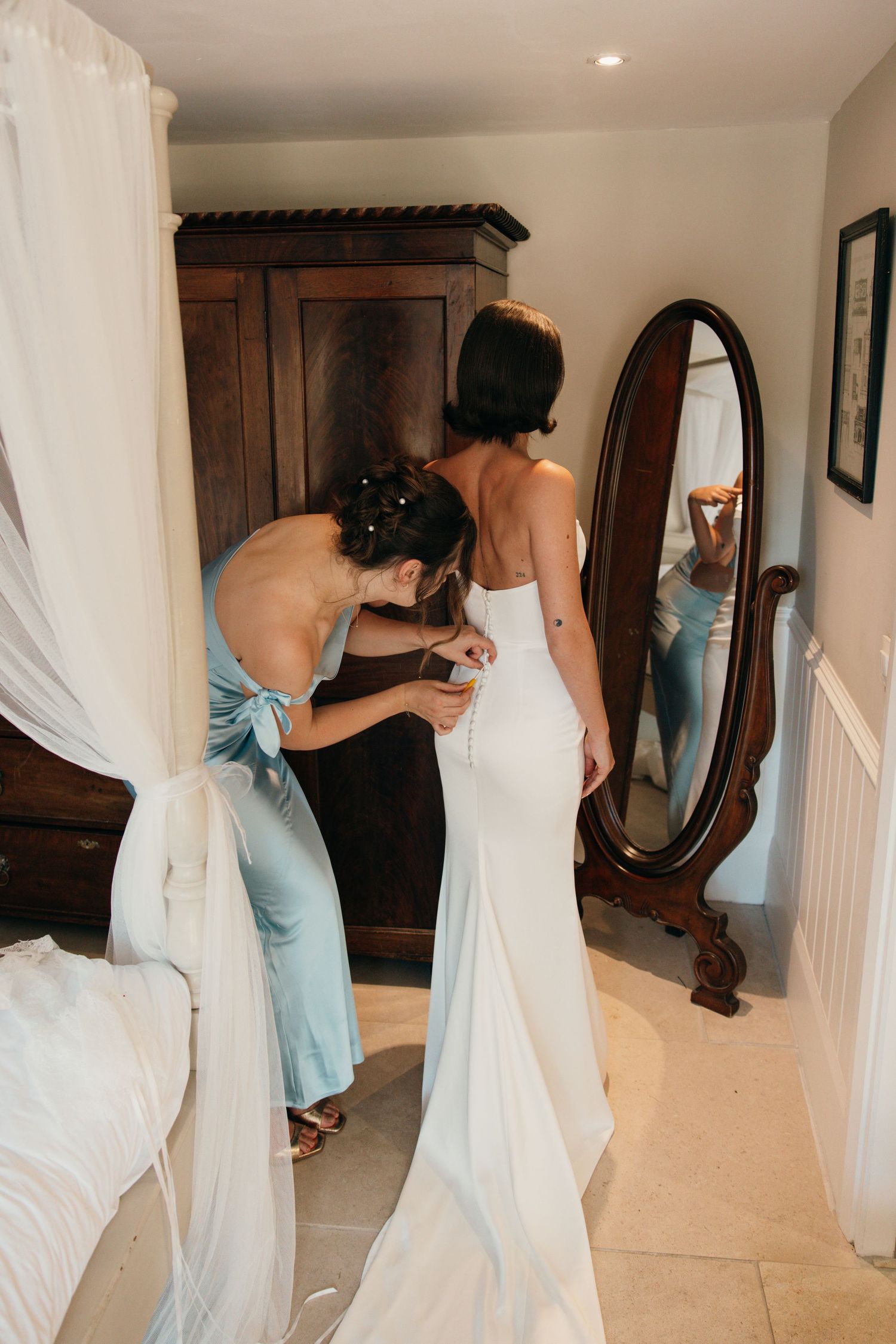 Bridesmaid helping bride button up white wedding dress while bride views reflection in vintage oval mirror.