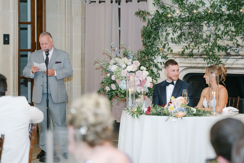 Man giving wedding toast at elegant reception while bride and groom smile at sweetheart table with floral arrangements.