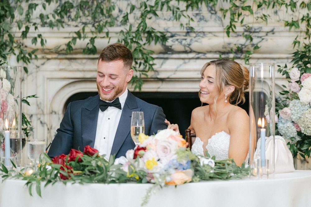Bride and groom laughing at sweetheart table with floral centerpieces and candlelight at elegant wedding reception.
