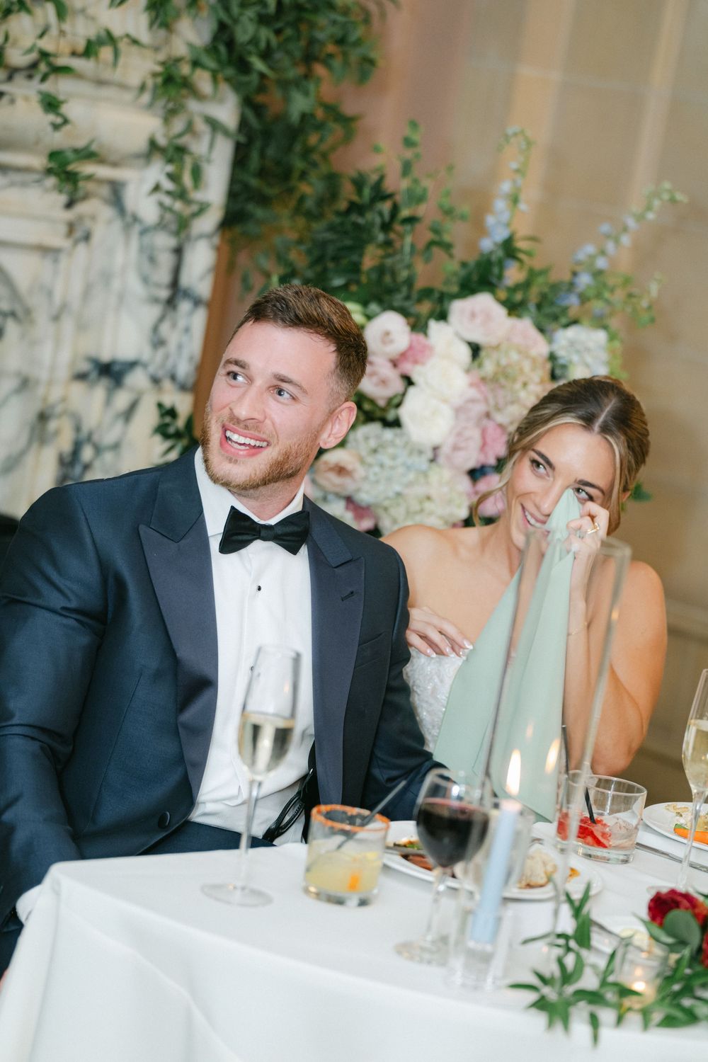 Bride and groom laughing at wedding reception table with champagne, floral arrangements, and candlelight in elegant venue.