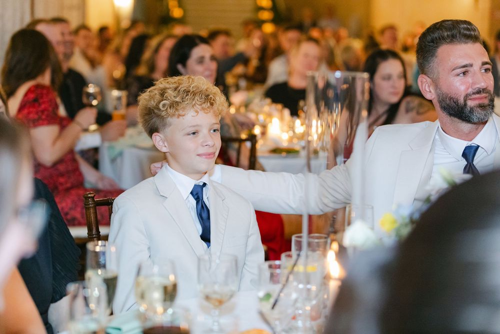 Boy in white suit with blue tie at a wedding reception dinner, seated at a candlelit table with guests.