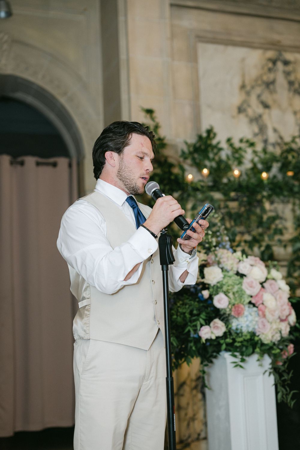 Man in beige suit giving a wedding speech at microphone stand with floral arrangements in background.