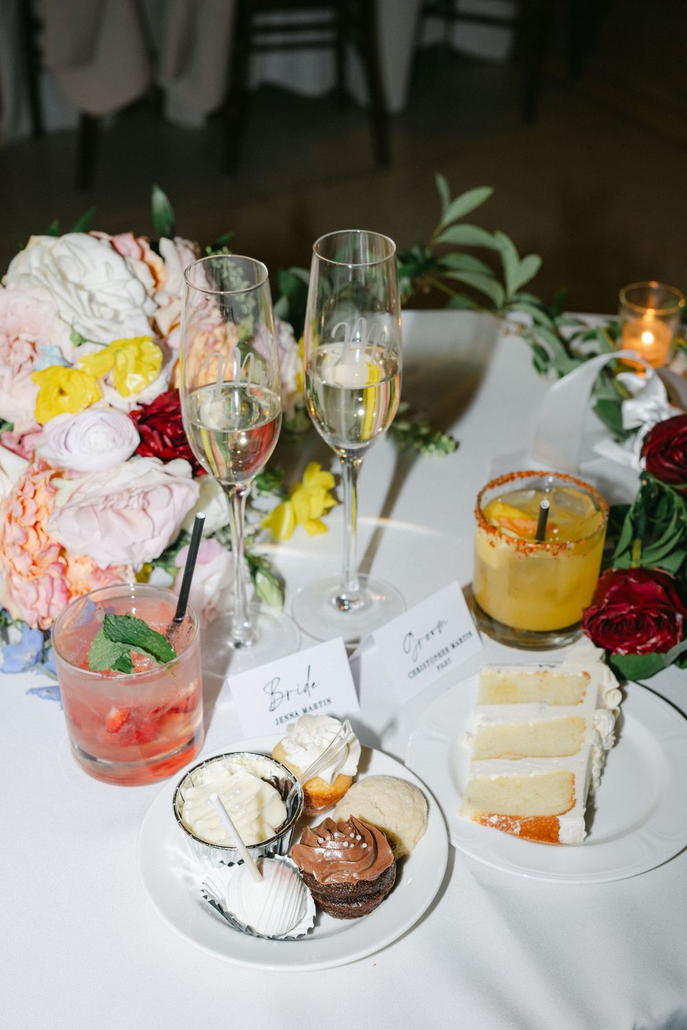 Wedding sweetheart table with champagne flutes, floral bouquet, dessert plate, and colorful cocktails on white linen.