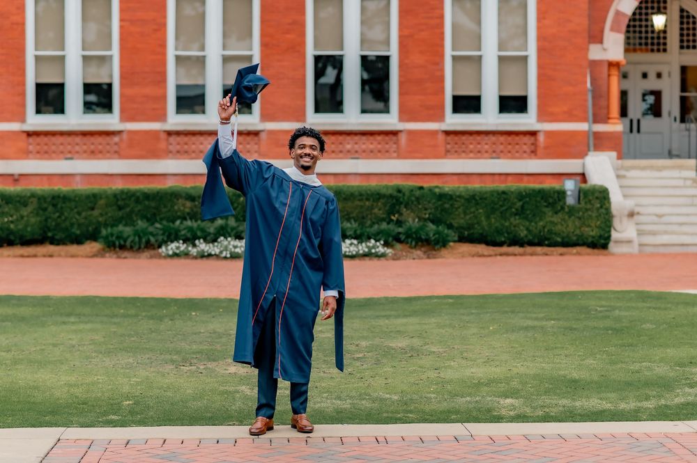 A smiling graduate in blue cap and gown raises his mortarboard outside a brick university building.