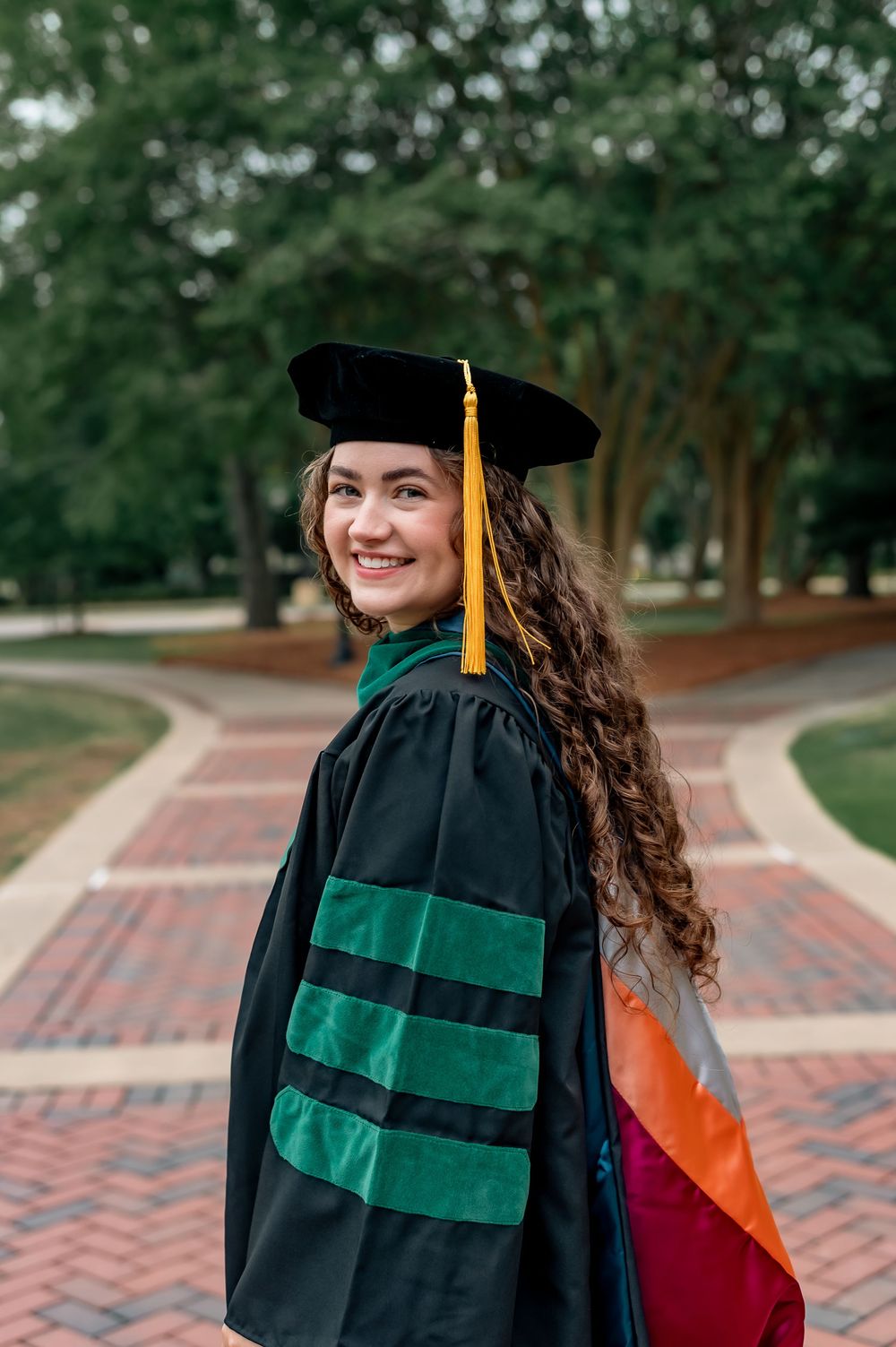 Smiling graduate in black doctoral gown with green velvet stripes and gold tassel cap on brick pathway.