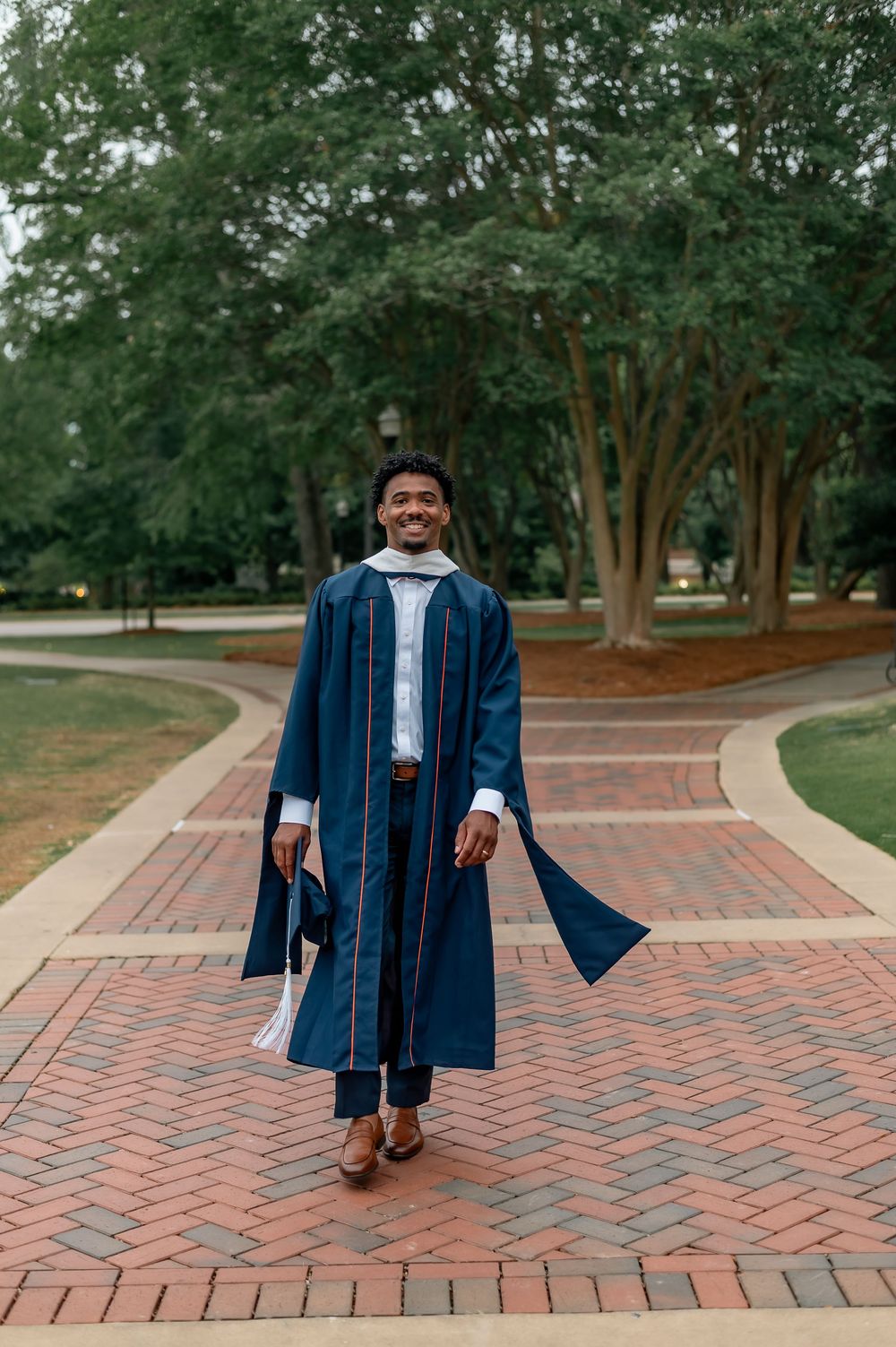 Smiling graduate in navy blue cap and gown walks along brick path surrounded by lush green trees.
