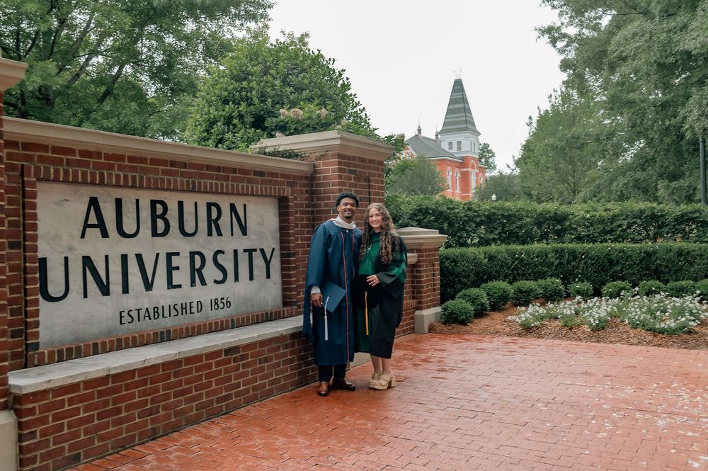 Two graduates in caps and gowns pose by the Auburn University brick entrance sign on a lush green campus.