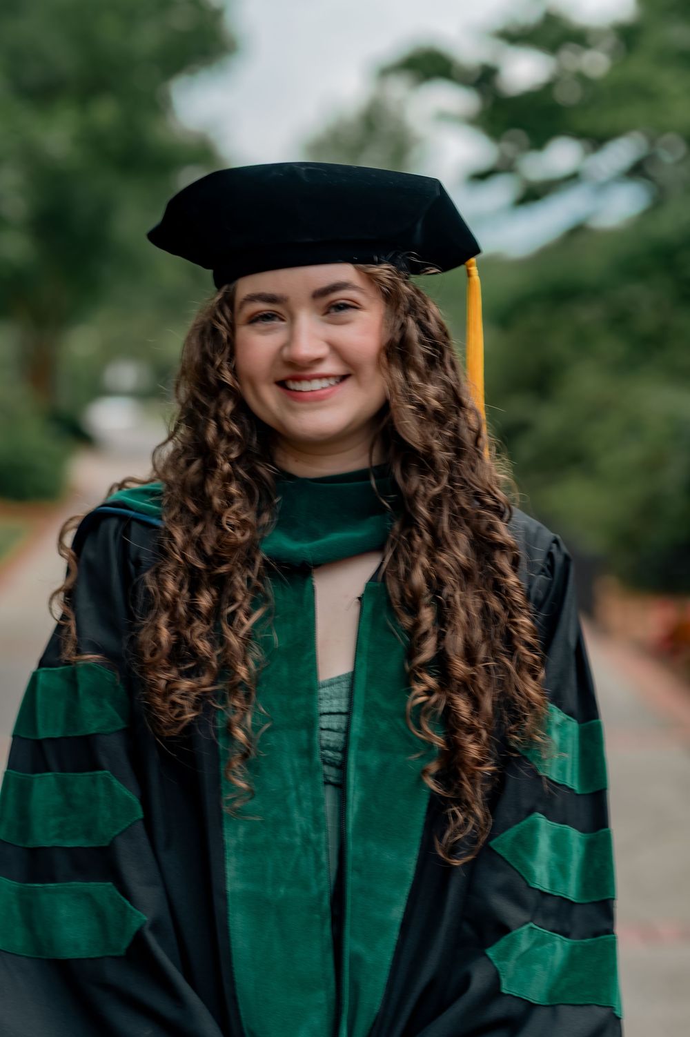 Smiling woman with curly hair wearing green and black doctoral graduation cap and gown outdoors.