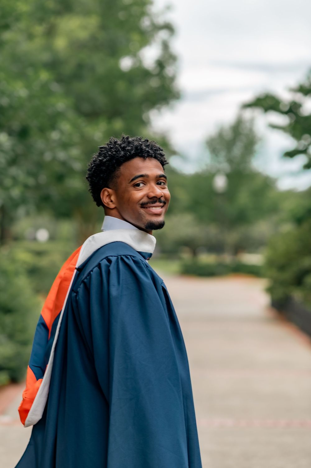 Smiling male graduate in navy blue and orange academic gown poses outdoors on a tree-lined campus path.