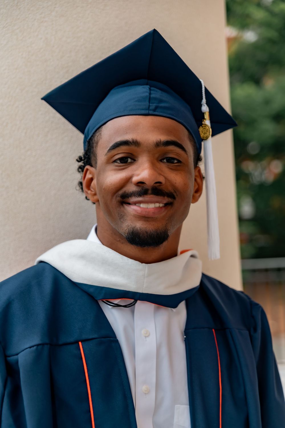 Smiling young man wearing navy blue graduation cap and gown with white hood posing outdoors.
