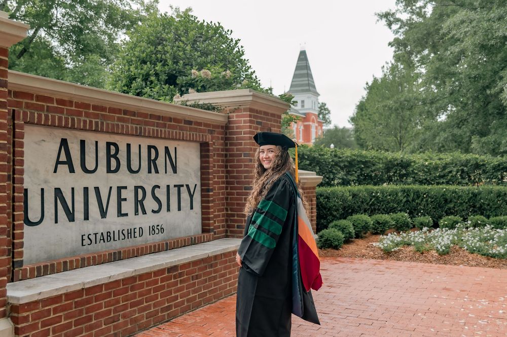 Graduate in cap and gown poses by Auburn University brick entrance sign on a cloudy day.