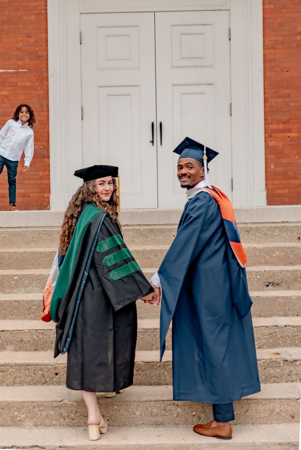 Two graduates in caps and gowns hold hands on steps outside a brick building, smiling back at the camera.