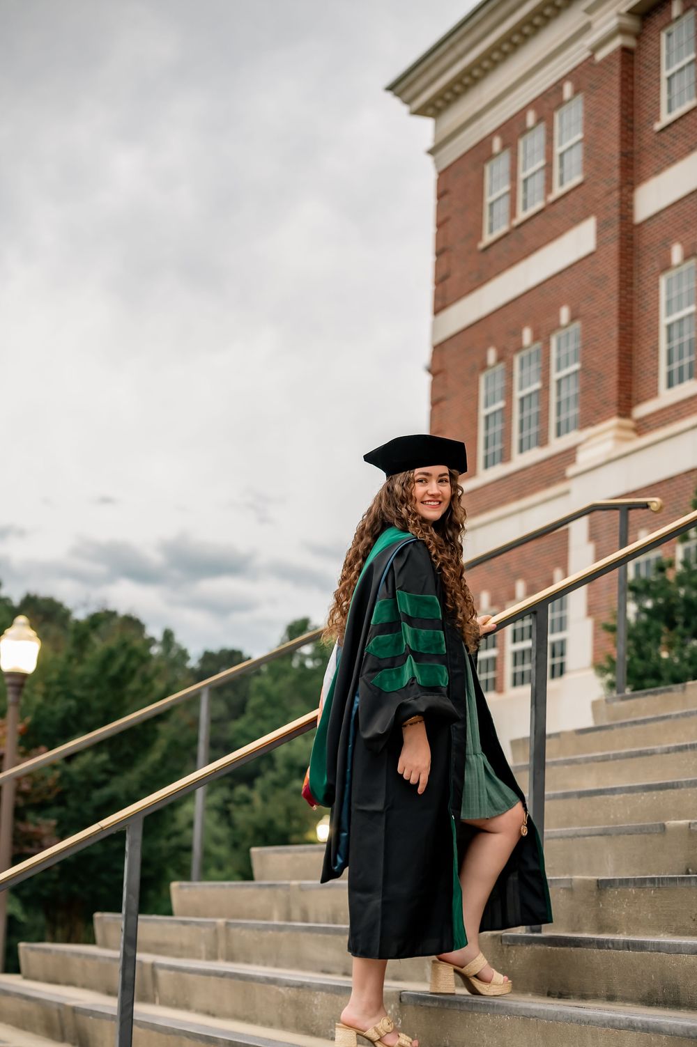 A woman in black and green doctoral regalia poses on steps outside a brick university building.