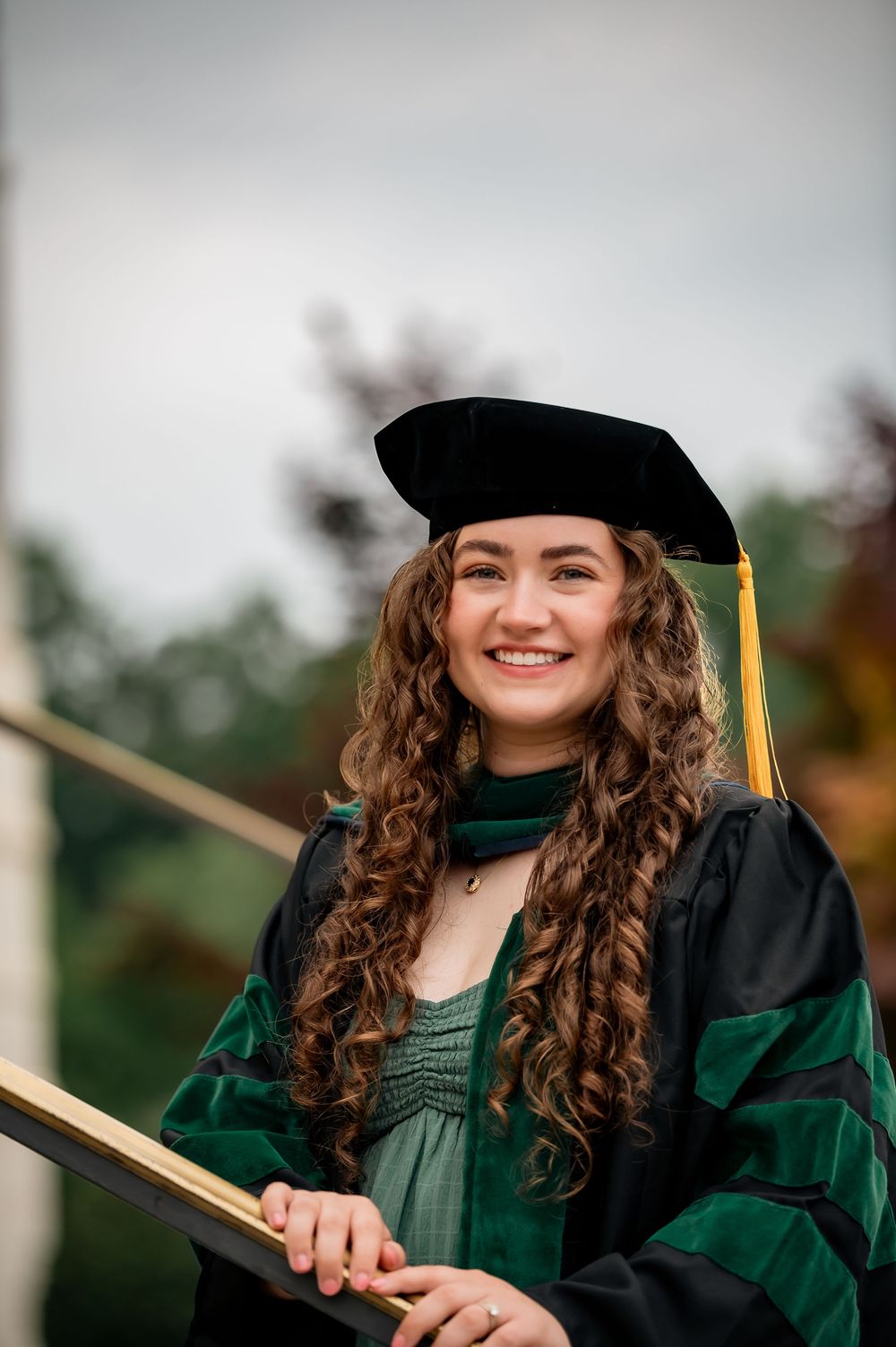 Smiling young woman in green and black doctoral graduation cap and gown holding a diploma outdoors.