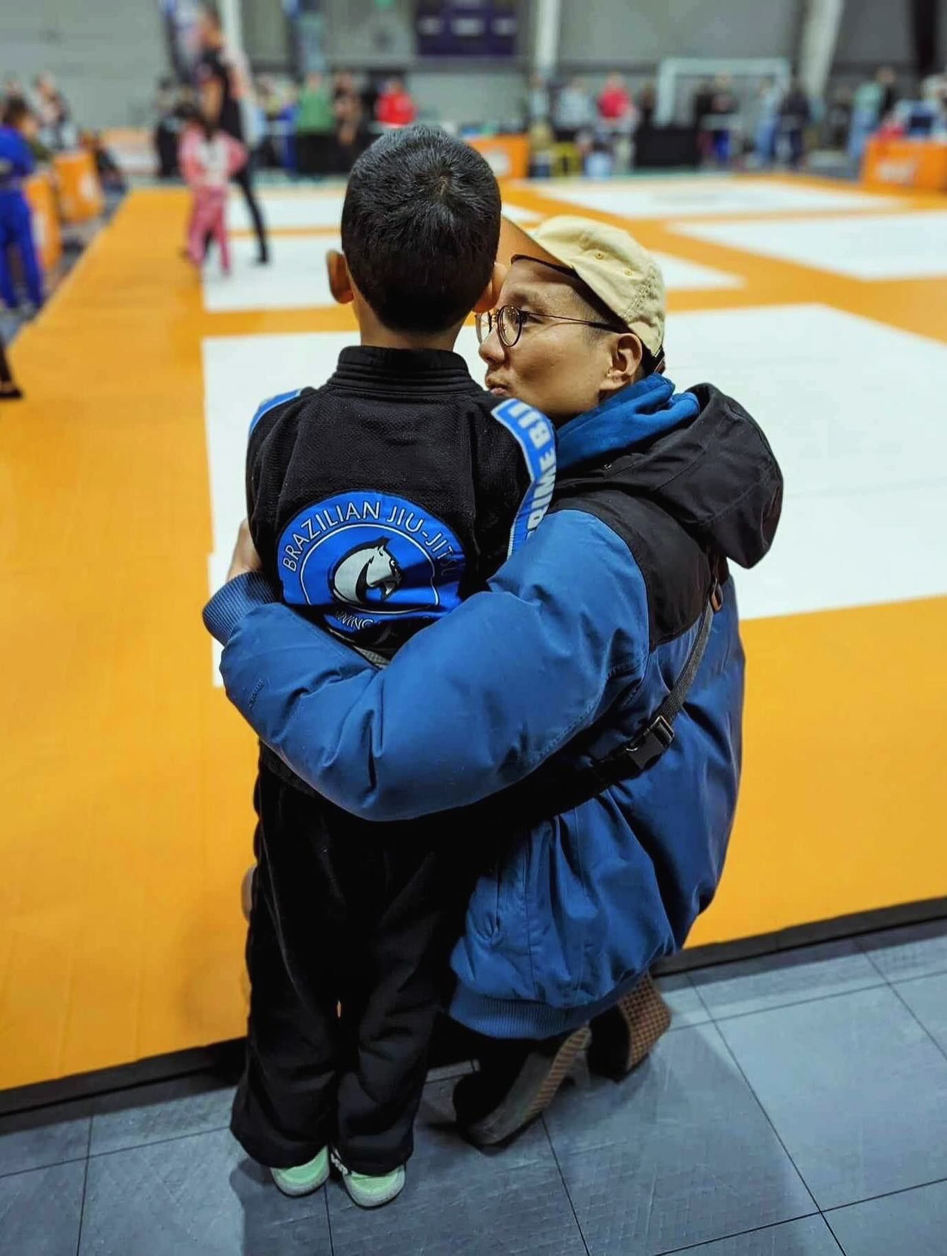 Adult kneeling to hug a child in a Brazilian Jiu-Jitsu uniform at an indoor martial arts tournament.
