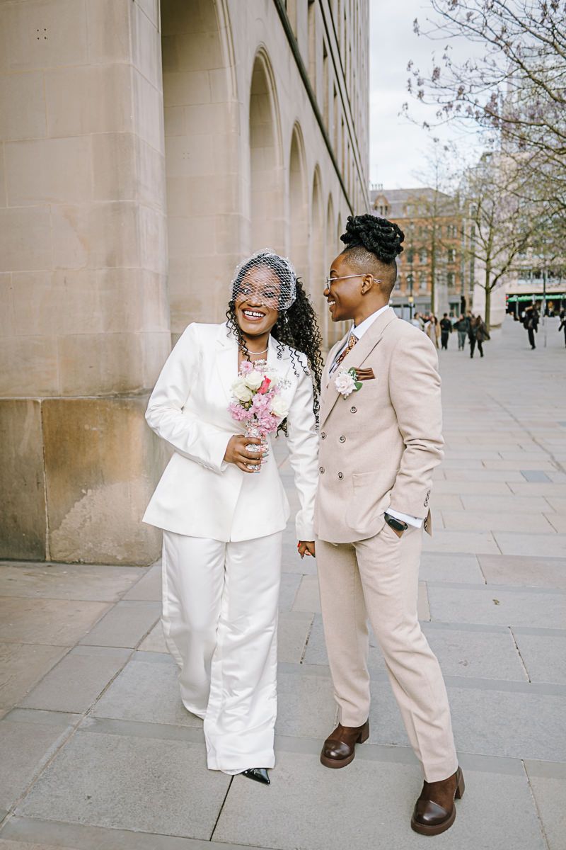 Two brides in elegant suits hold hands and laugh together outside a stone building on their wedding day.