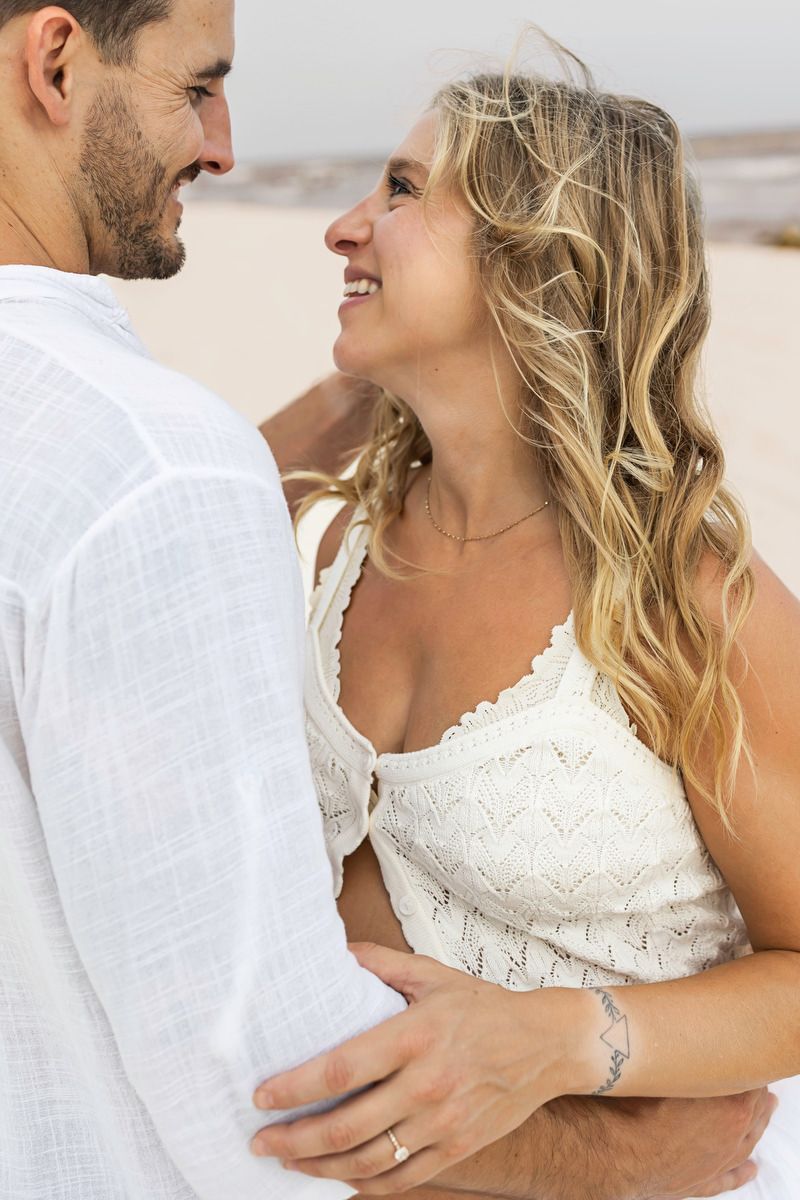 A smiling couple in white outfits embrace on a sandy beach, gazing lovingly at each other.