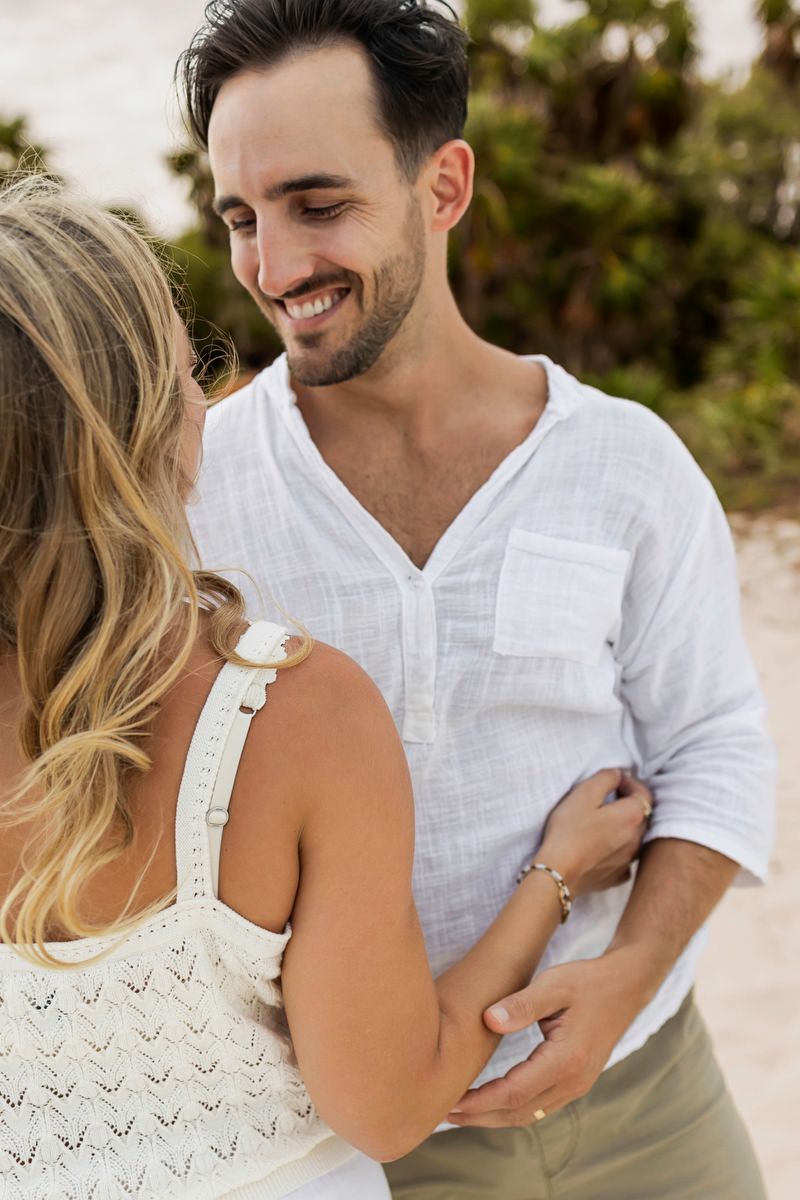 A smiling man in a white linen shirt embraces a blonde woman in a white dress outdoors.