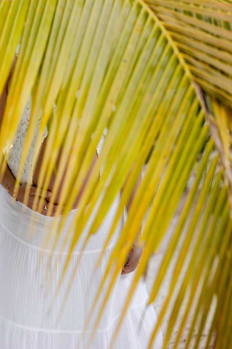 Person in white dress partially hidden behind a large tropical palm leaf with yellow and green fronds.