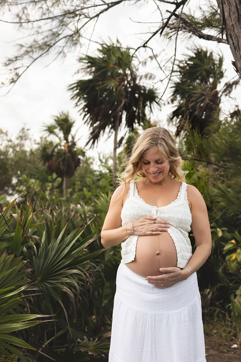 Smiling pregnant woman in white dress cradling her baby bump outdoors among tropical palms.