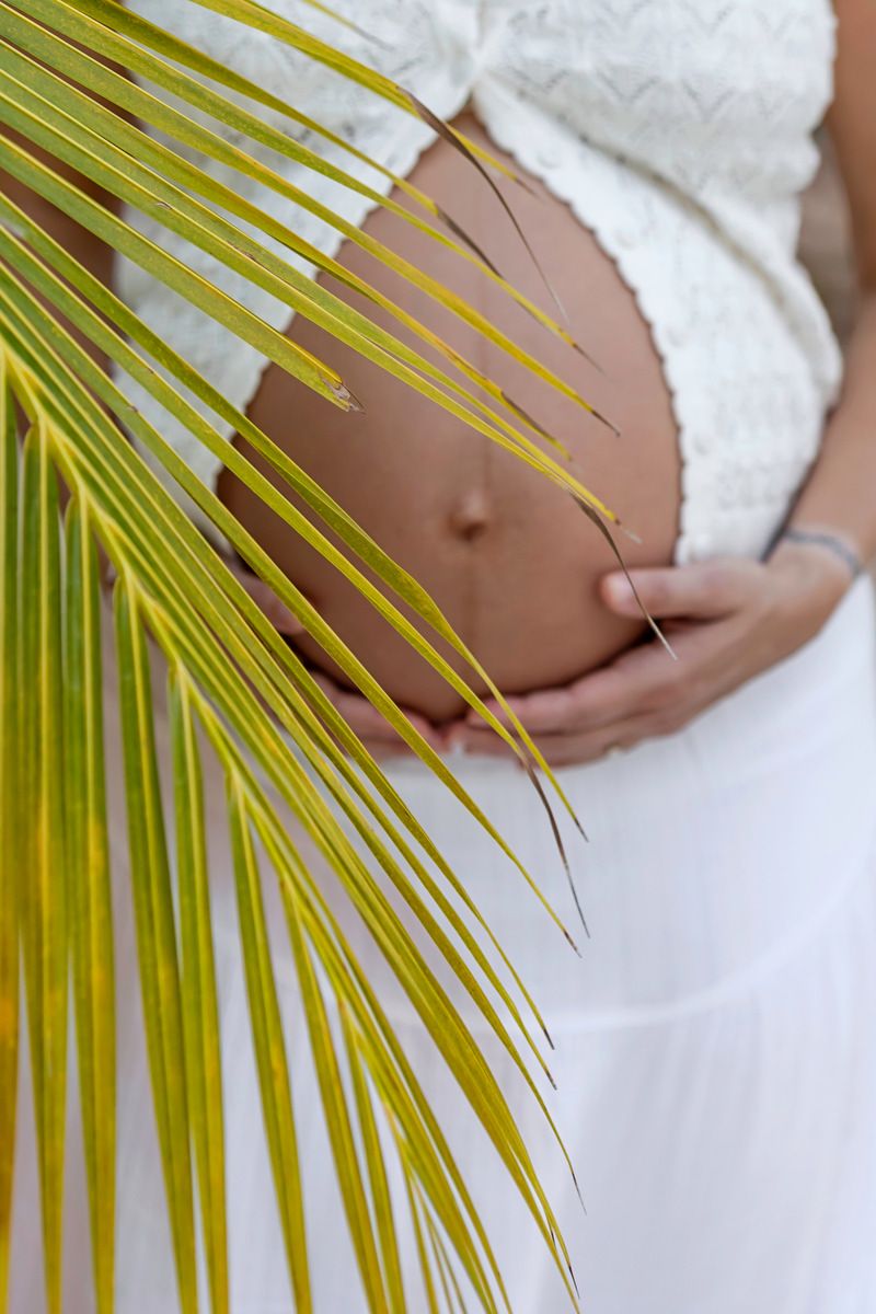 Pregnant woman in white dress cradling her baby bump behind a tropical palm leaf outdoors.