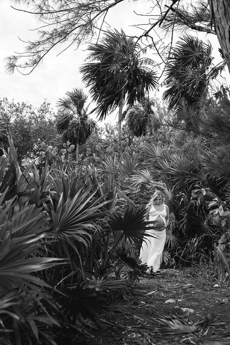 Black and white photo of a woman in a white dress standing among lush tropical palm plants and trees.