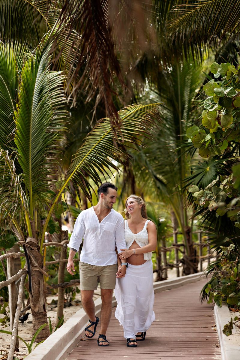 Couple walking hand-in-hand on a wooden boardwalk path surrounded by lush tropical palm trees at a beach resort.