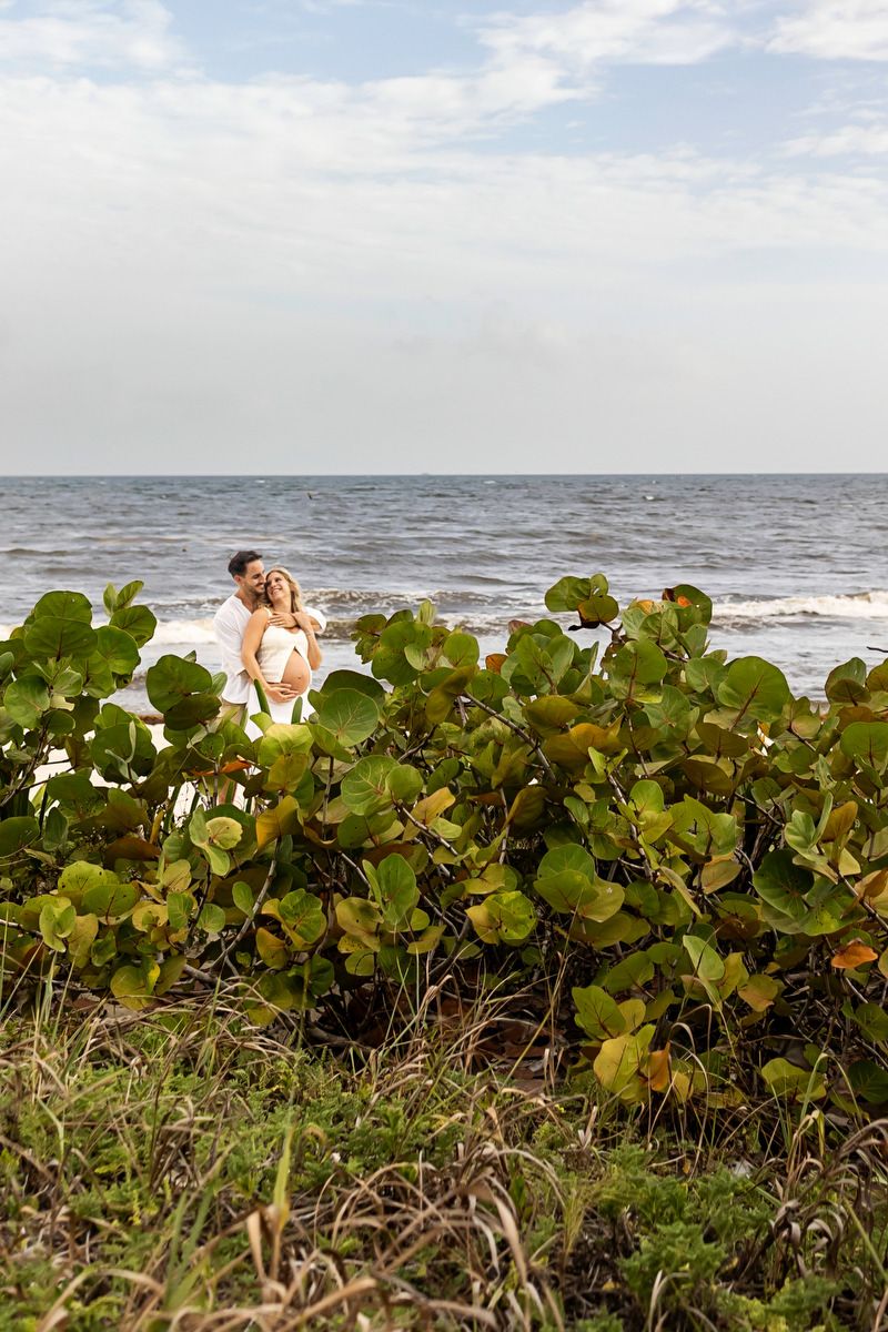 Couple embracing on a tropical beach with lush green sea grape plants in the foreground and ocean waves behind them.
