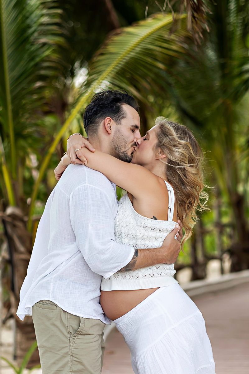 Pregnant couple kissing romantically outdoors surrounded by lush tropical palm trees, both dressed in white.