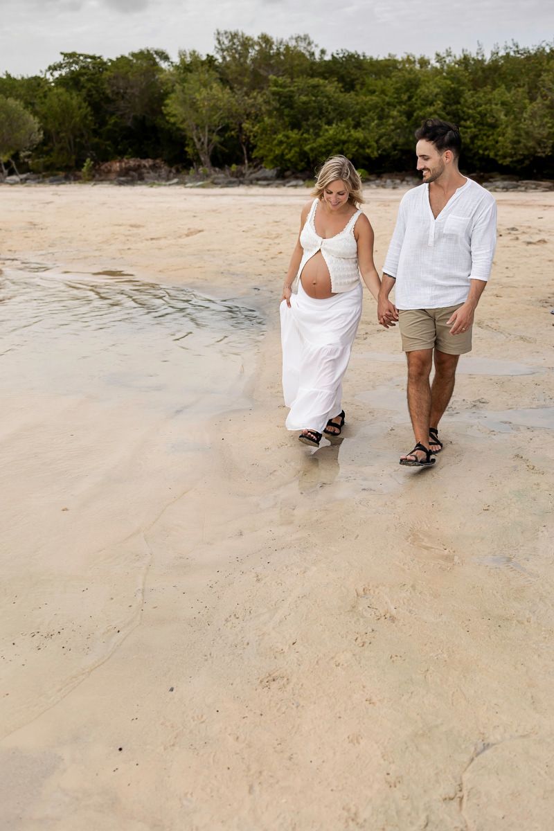 Pregnant couple holding hands walking on sandy beach shore, woman in white dress, man in white shirt and khaki shorts.