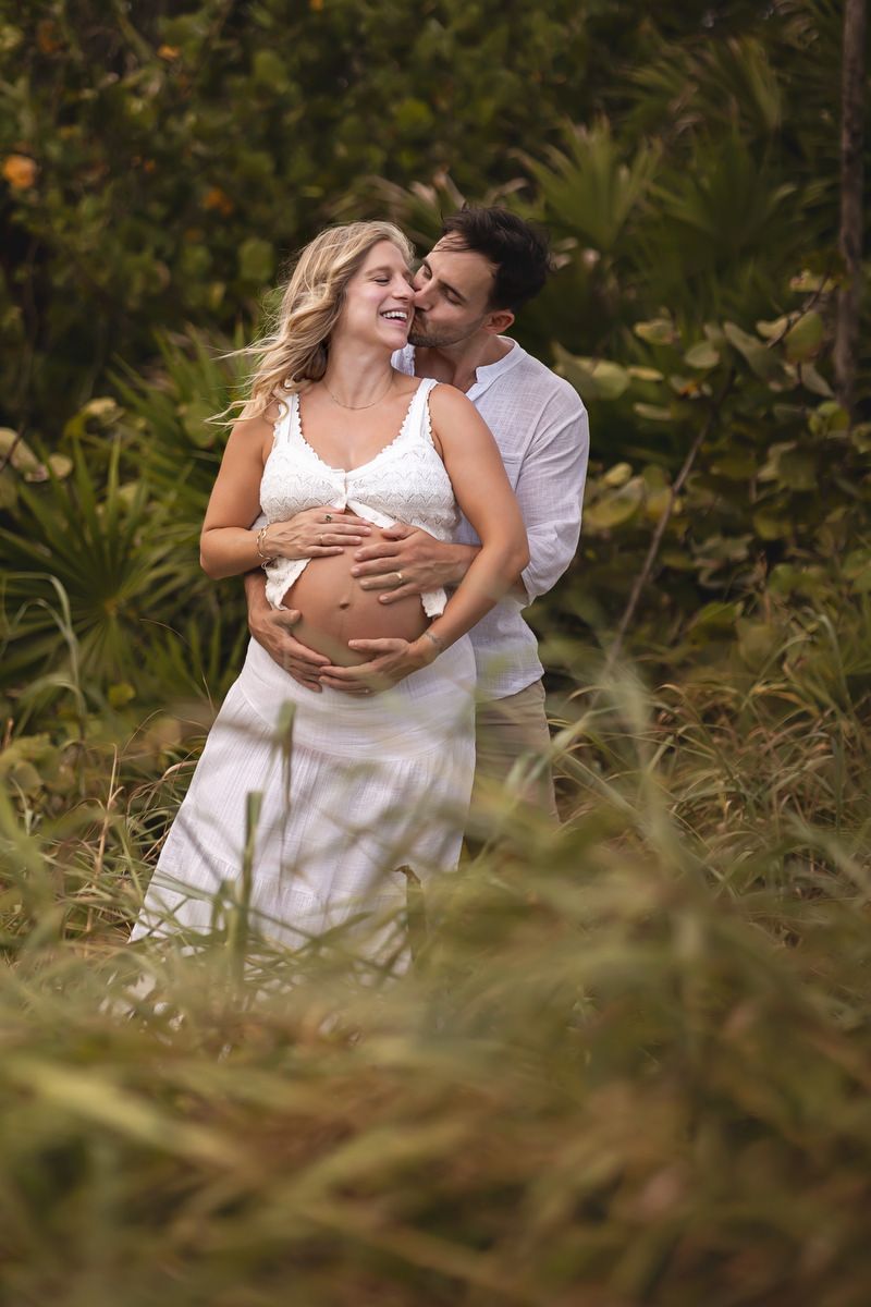 A couple shares a tender moment outdoors, man kissing pregnant woman's cheek while cradling her baby bump.