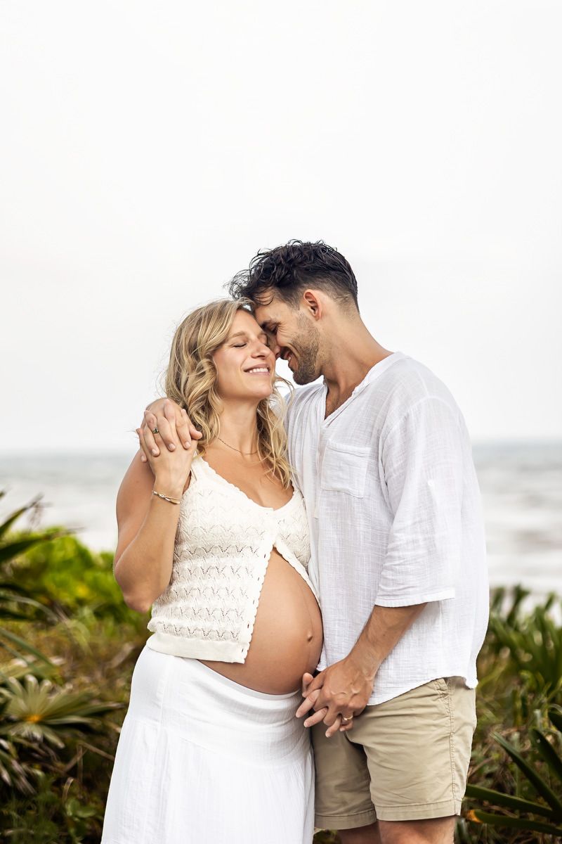 Pregnant couple embracing on a beach, woman in white crop top and skirt, man in white shirt and khaki shorts.