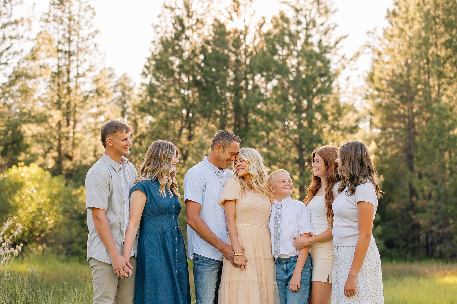 Family of six posing together in a sunlit meadow surrounded by tall pine trees during a golden hour outdoor photo session.