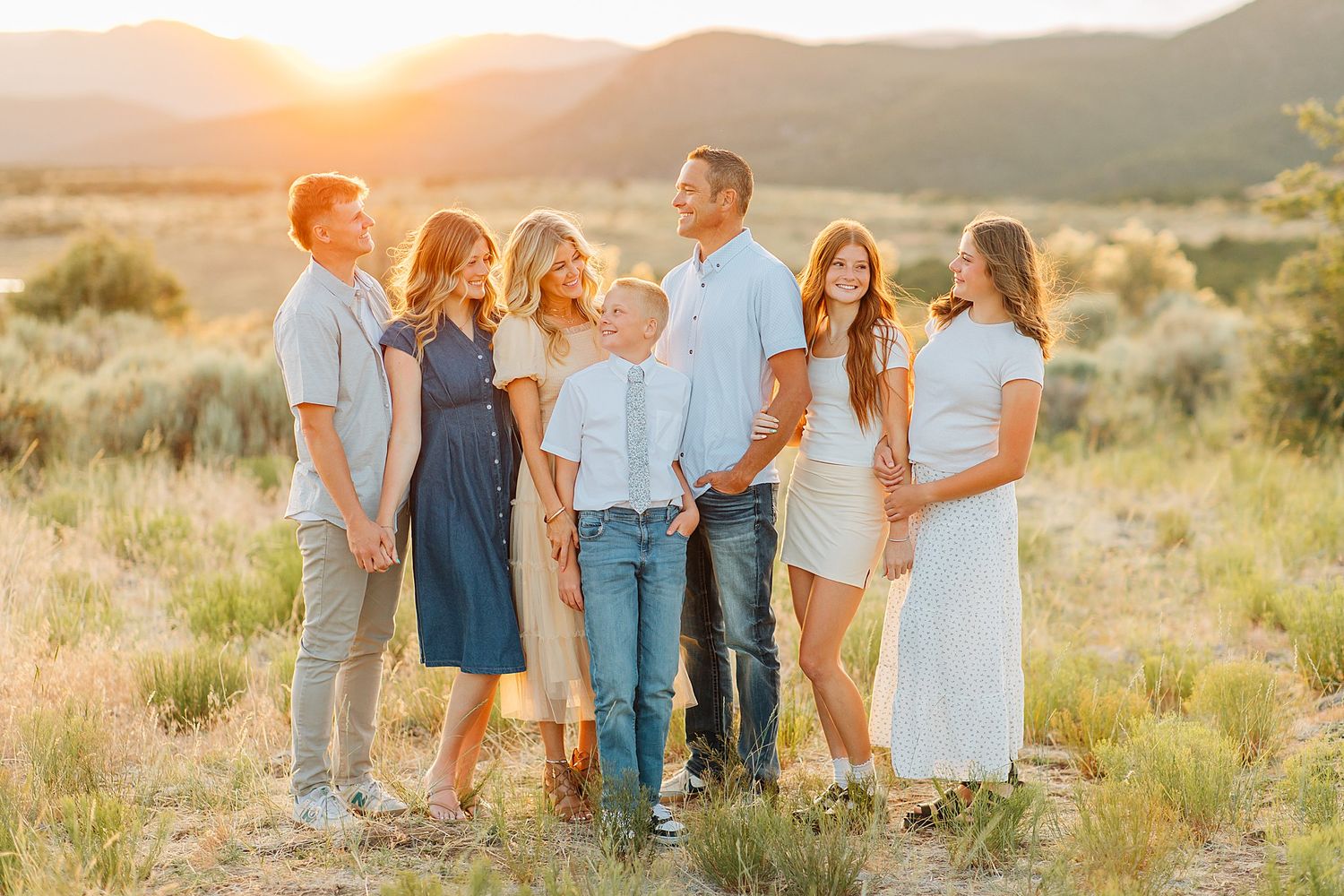 Family of six posing outdoors at golden hour with mountain landscape and green fields in background.