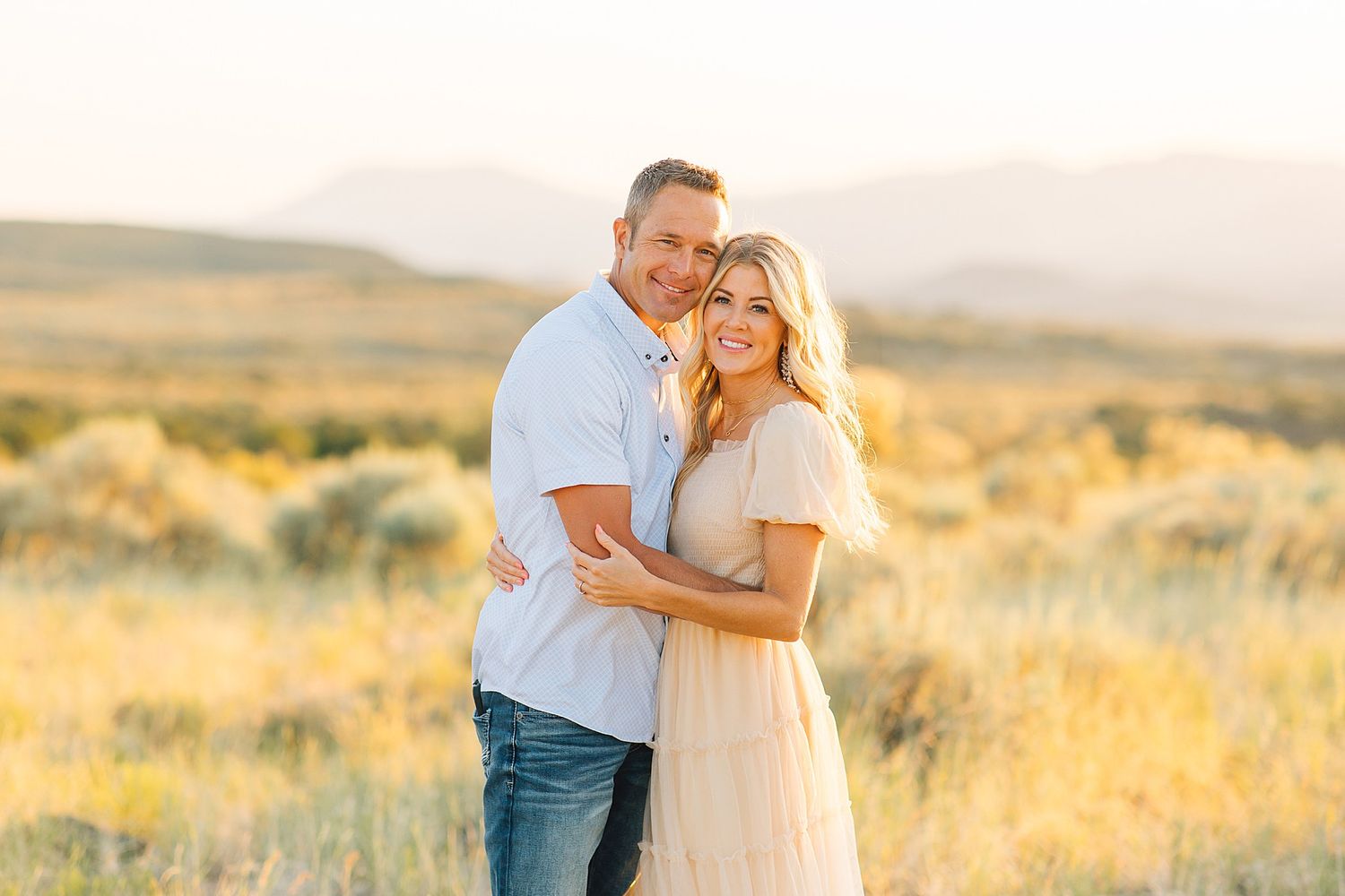 Couple embracing in golden field with mountain backdrop during sunset engagement photo session.