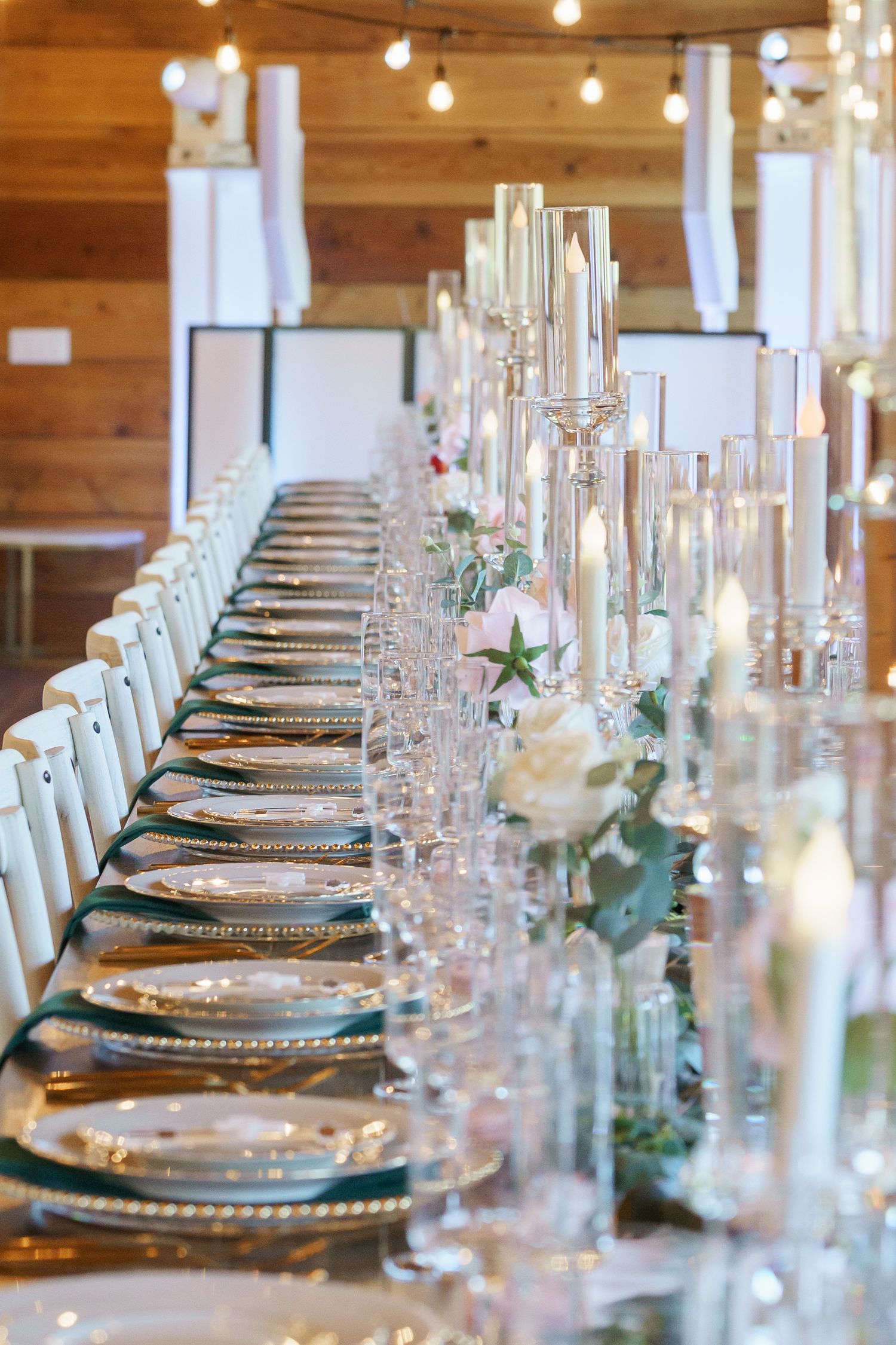 Elegant wedding reception table set with crystal glasses, white roses, tall candles, and gold charger plates in a rustic barn venue.