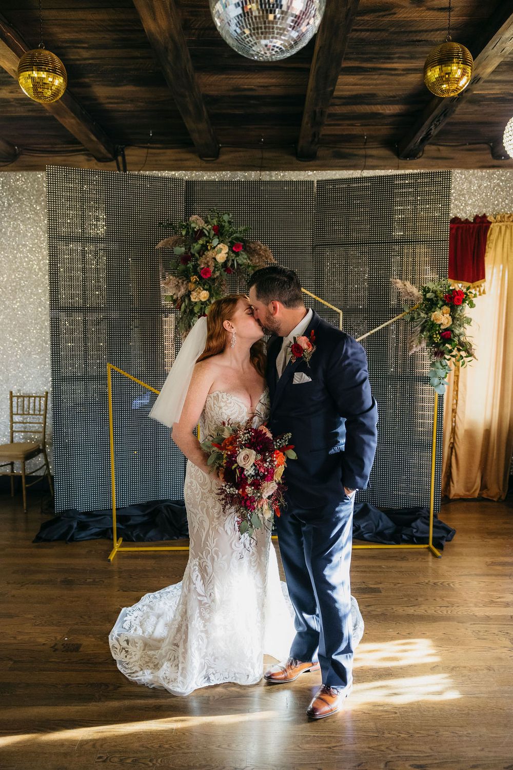Bride and groom share a kiss at indoor wedding ceremony with floral arch backdrop and disco ball at Arcada Theater in Saint Charles, IL