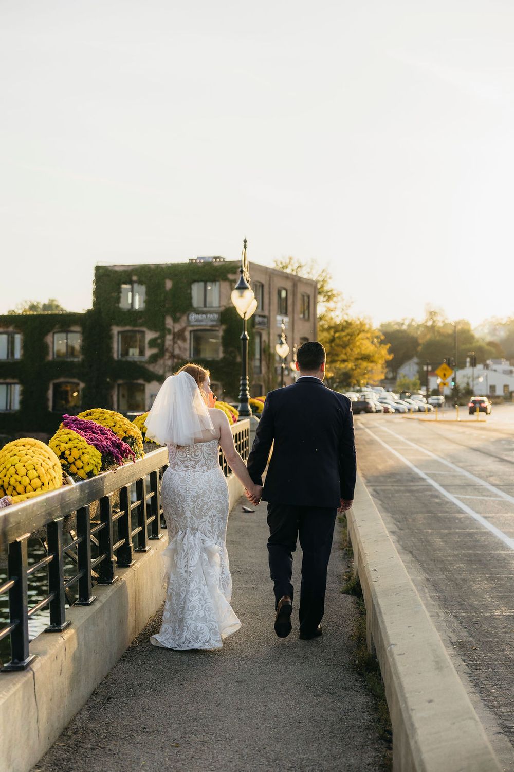 Bride and groom walk hand in hand along a scenic waterfront path lined with colorful fall flowers in Saint Charles, IL