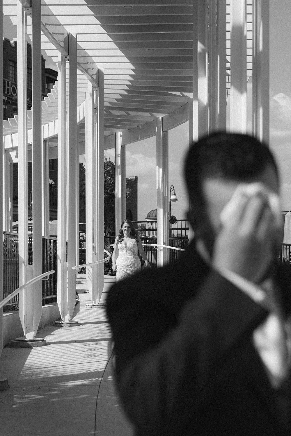 Black and white photo of groom wiping tears as bride walks toward him down a sunlit corridor during wedding first look in Saint Charles, IL