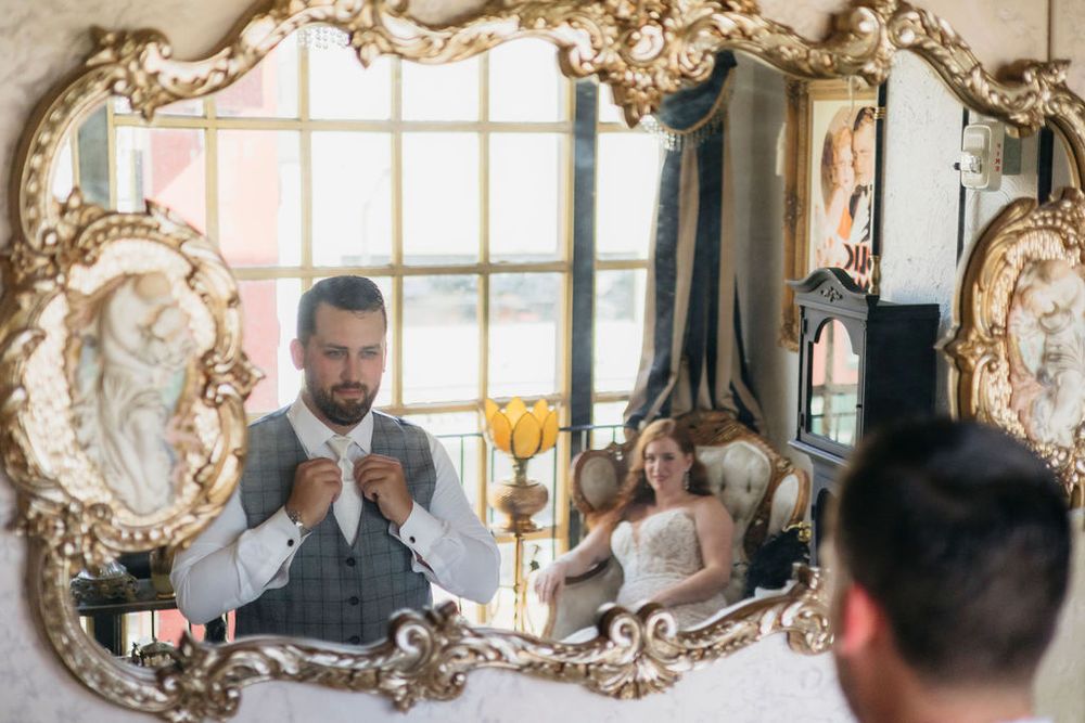 Groom adjusting tie reflected in mirror, bride in wedding dress seated in elegant room background at Arcada Theater in Saint Charles, IL