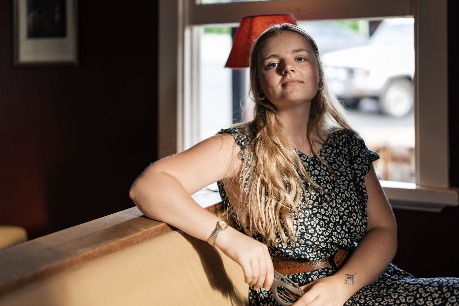 A young blonde woman in a floral dress leans on a windowsill in a casual restaurant setting.

Back Bar