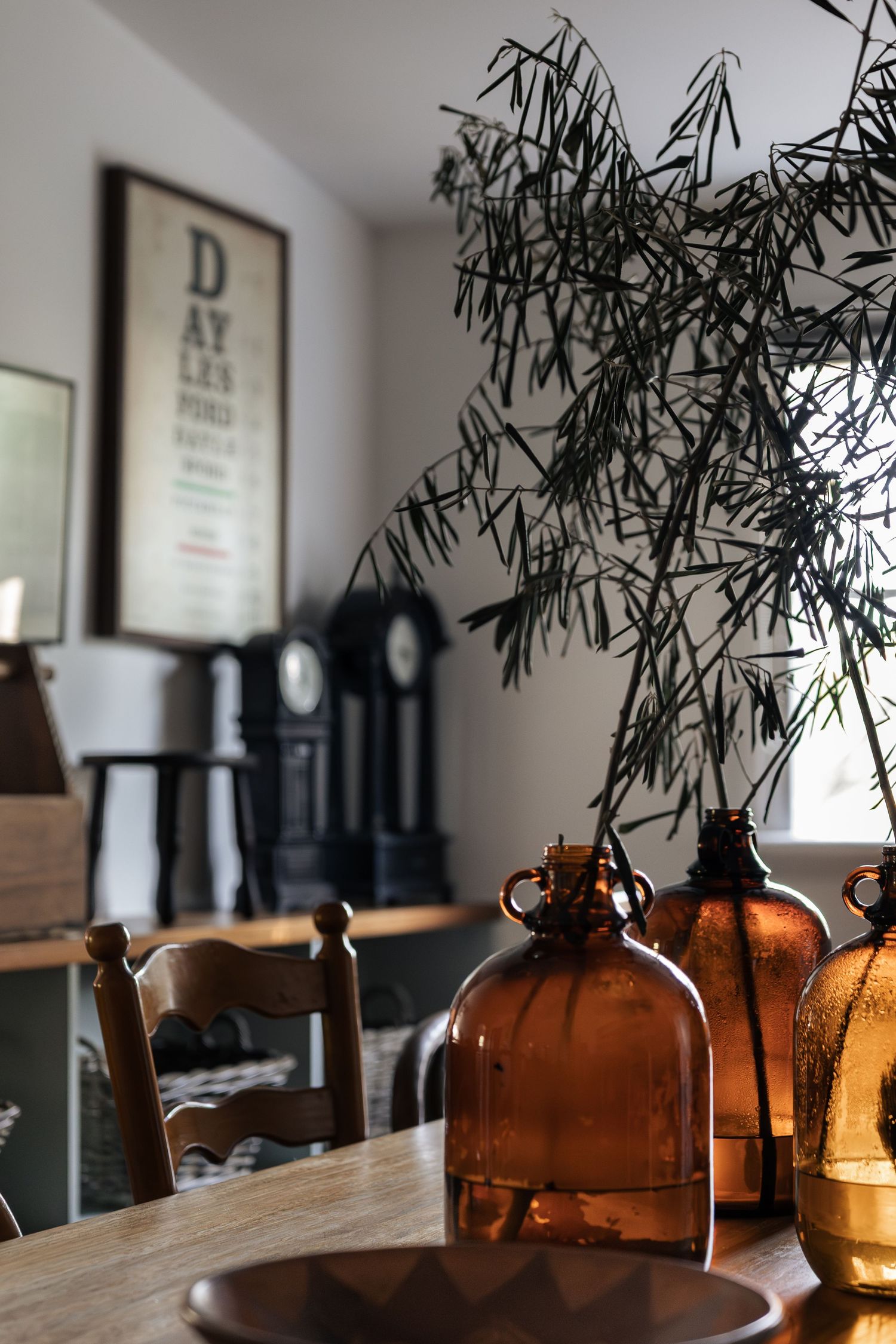 Rustic dining table with amber glass bottles, green botanical stems, wooden chairs, and vintage decor in background.

Piccadilly House