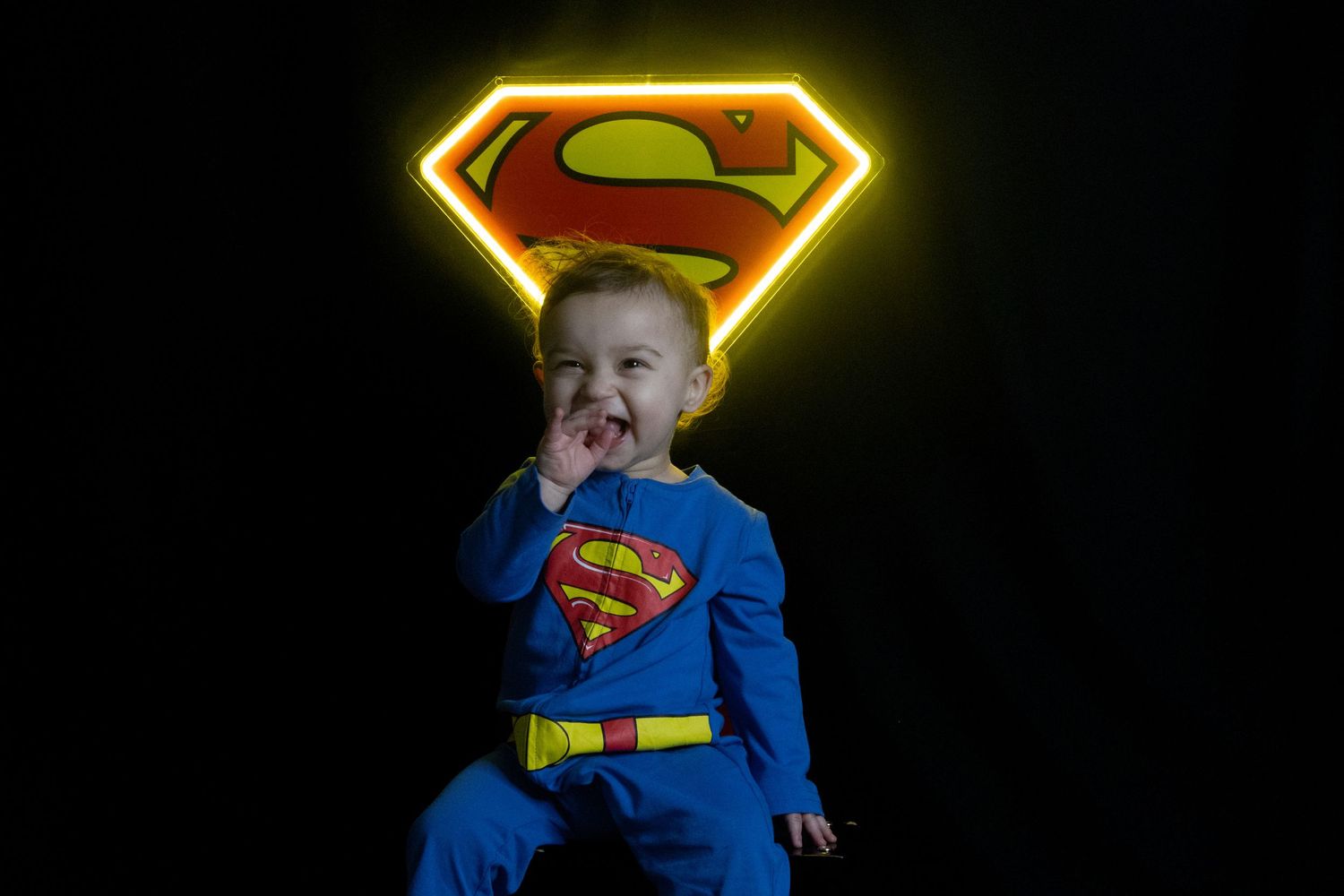 Baby in Superman costume sits before glowing Superman logo on black background, smiling with hand near mouth.