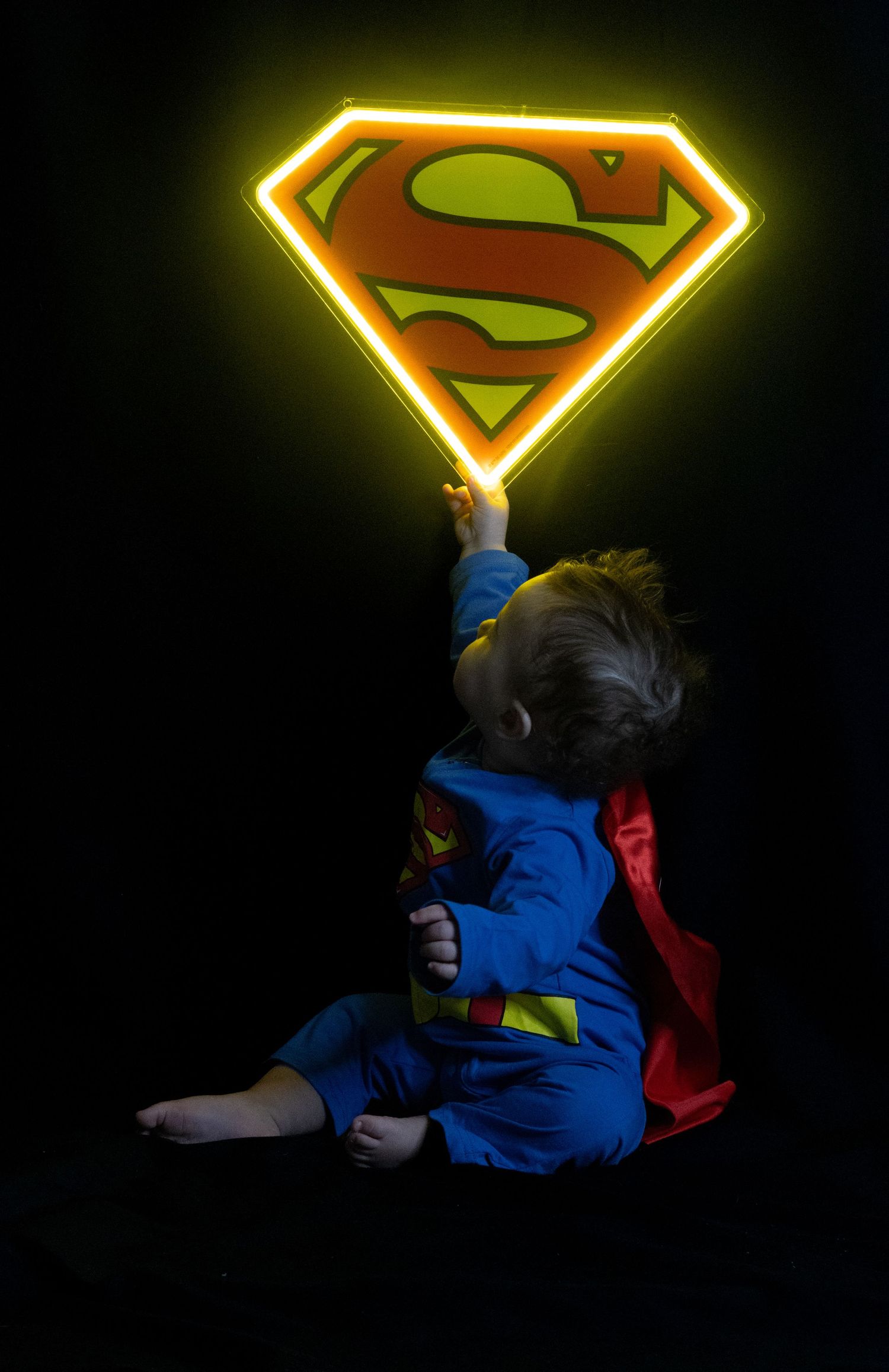 A baby dressed in a Superman costume reaches up to touch a glowing neon Superman logo sign against a dark background.