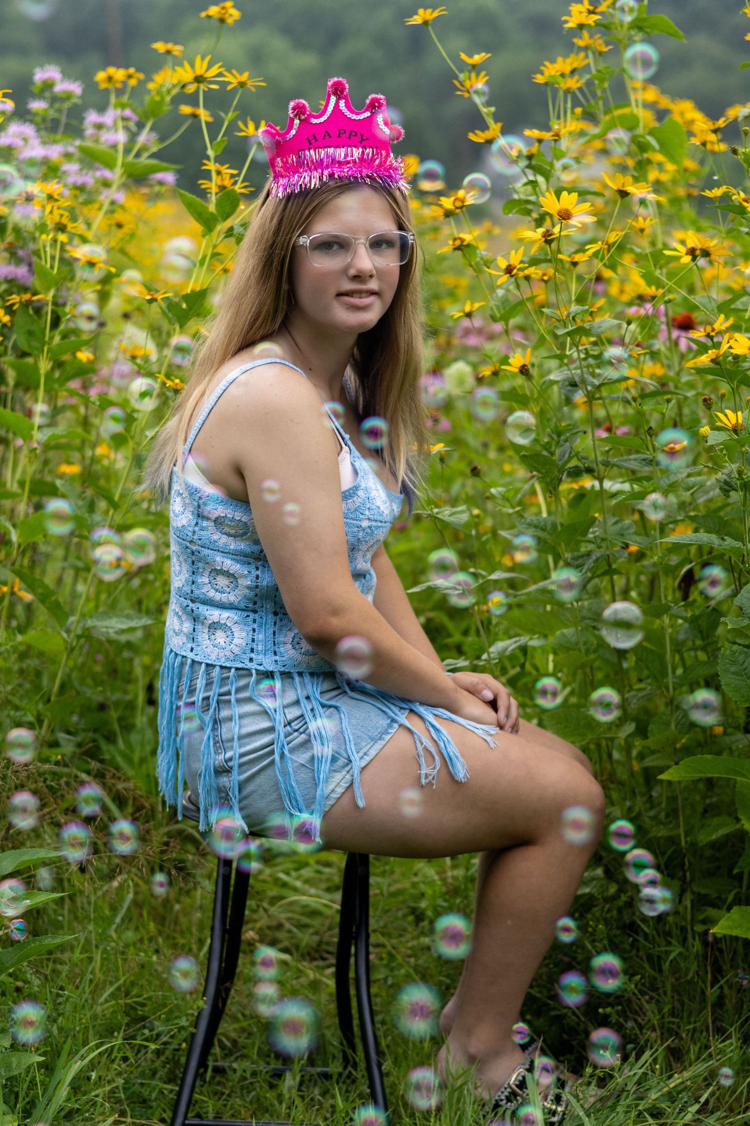 Young woman in blue crochet dress and pink crown sits on chair surrounded by wildflowers with bubbles floating around her.