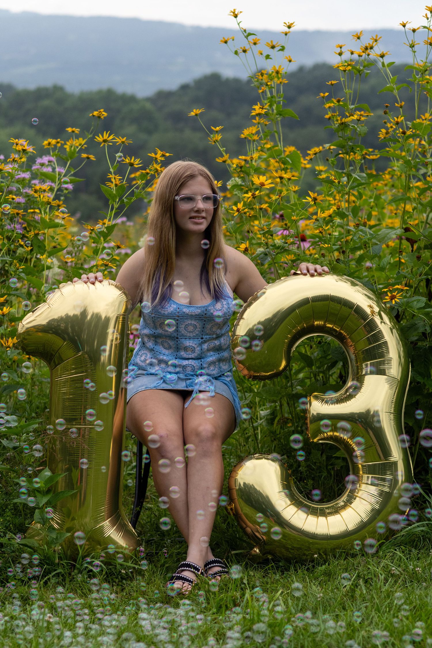 Teen girl sitting among yellow wildflowers holding gold number 13 balloons, surrounded by soap bubbles outdoors.