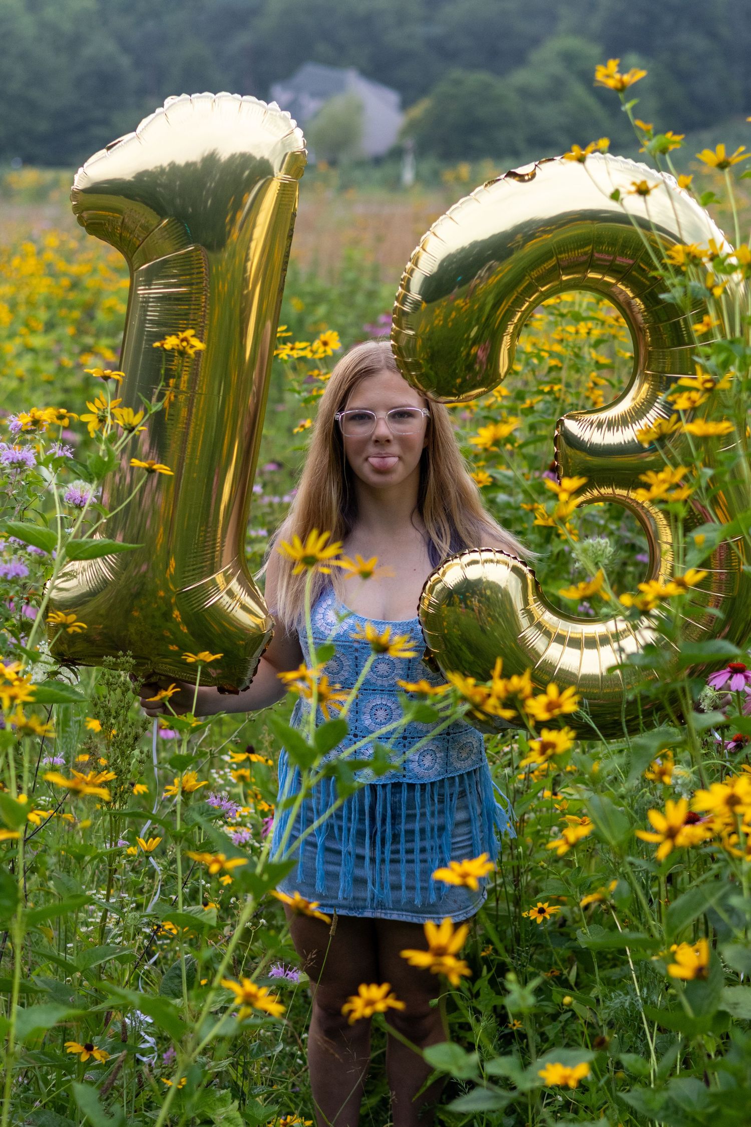Teen girl holding gold number 13 balloons in a field of yellow wildflowers, celebrating her 13th birthday.